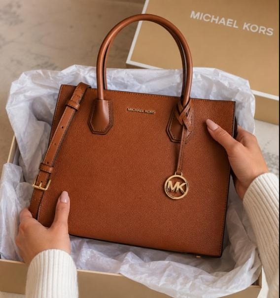 Hands lifting a cognac Michael Kors Mercer leather tote from its signature box with tissue paper