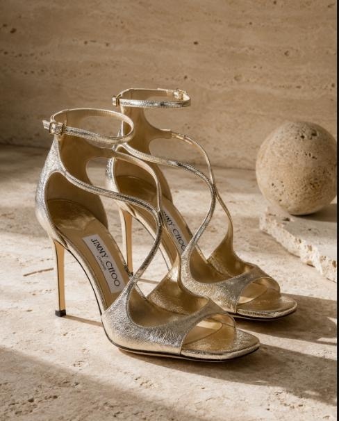 Pair of Jimmy Choo metallic gold strappy stiletto sandals on a sunlit stone surface