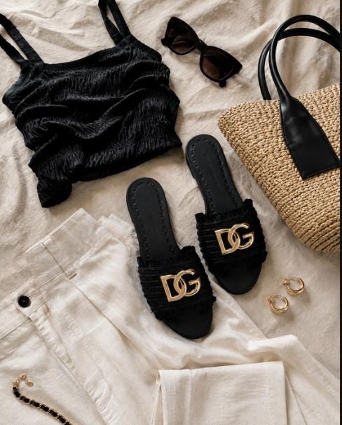 Flat-lay of black Dolce & Gabbana DG-logo mule slides styled with a black textured top, raffia tote, and gold hoops