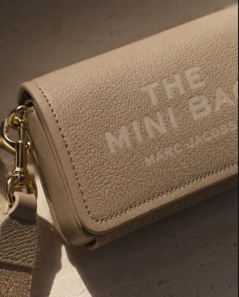 Close-up of tan pebbled leather Marc Jacobs "The Mini Bag" crossbody with gold clasp hardware in direct sunlight